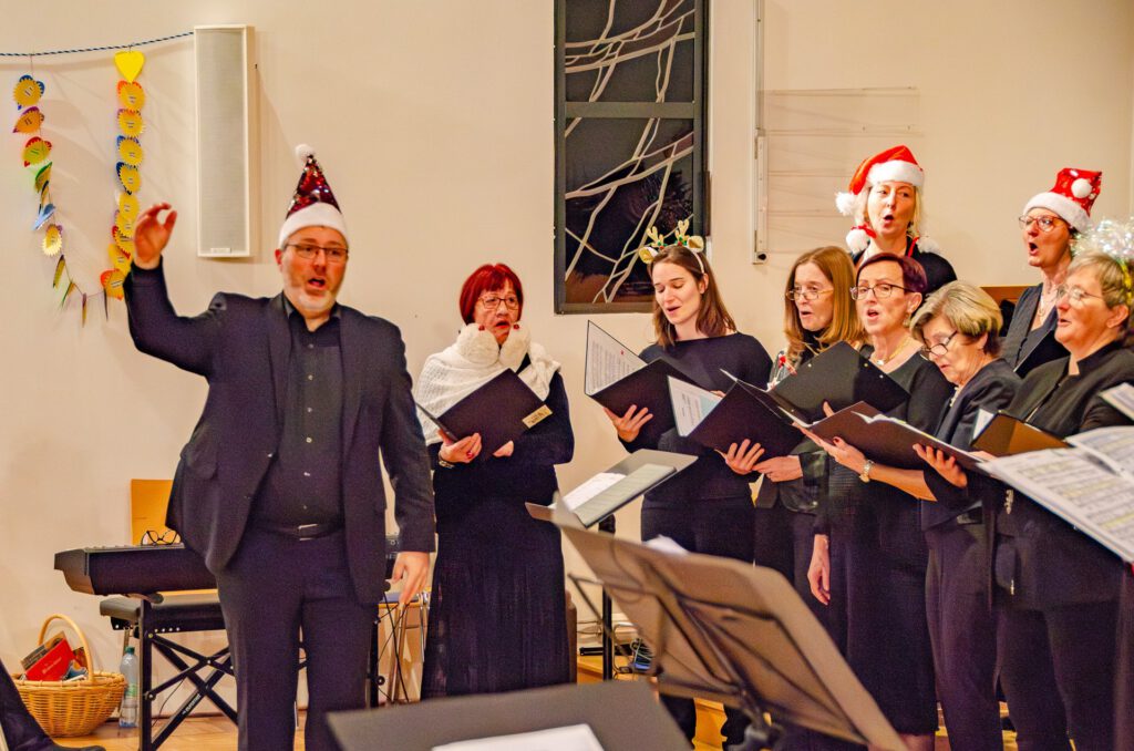 Der Chorleiter dirigiert mit Weihnachtsmütze, Sängerinnen singen aus ihren Notenmappen, einige ebenfalls mit Mützen und Rentiergeweih
