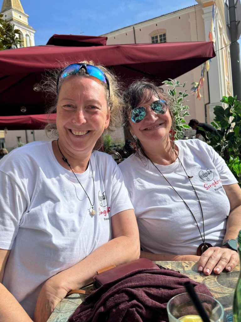 Zwei Sängerinnen in Singkreis-T-Shirts lachen in einem Straßencafé in Poreč.