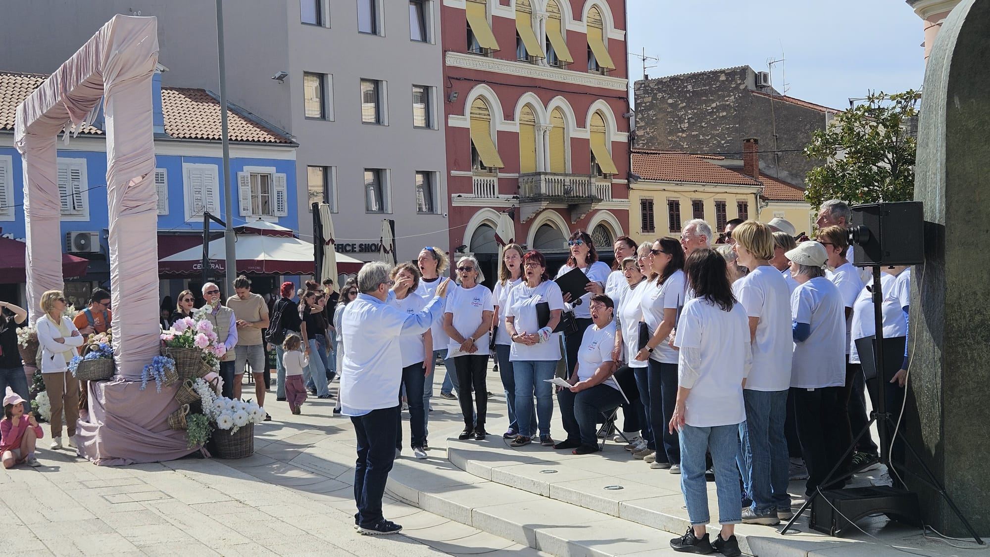 Der Singkreis Raaba singt in weißen Chor-T-Shirts auf einem Platz in der Altstadt von Poreč, Passanten hören zu.