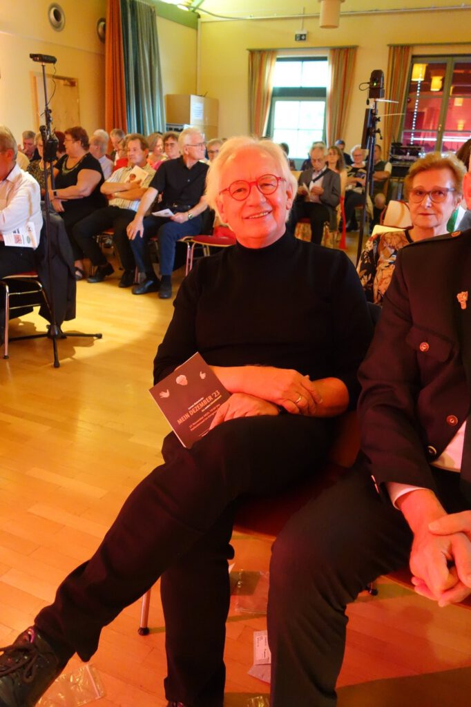 Lorenz Maierhofer mit weißem Haar und roter Brille sitzt lächelnd in der ersten Reihe, hinter ihm das Publikum im Saal