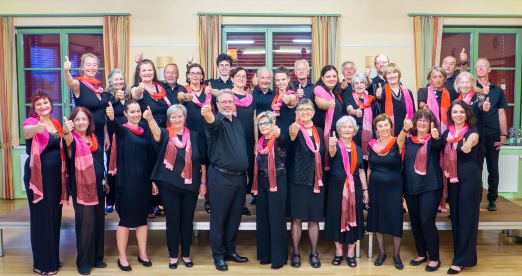Gruppenfoto des Singkreis Raaba in Schwarz mit pinken und roten Schals, alle zeigen den Daumen nach oben, der Chorleiter steht vorne in der Mitte