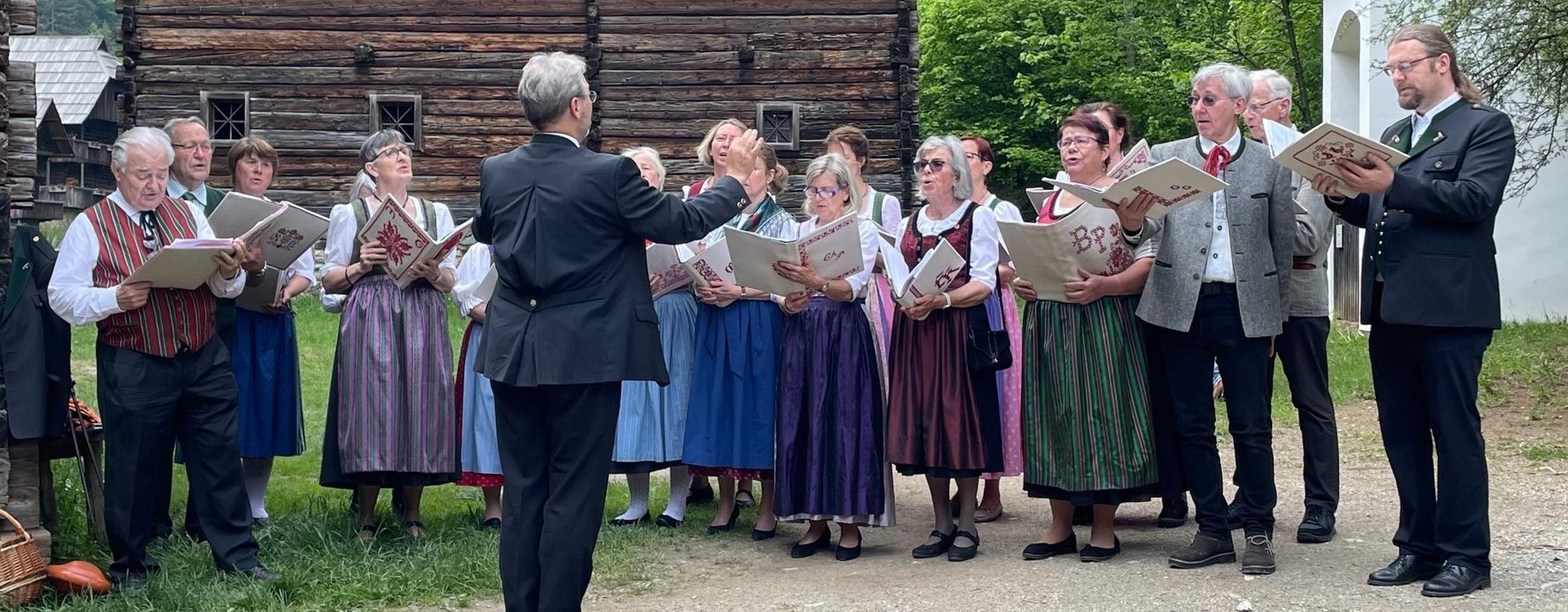 Sängerinnen und Sänger in Tracht singen aus Notenmappen vor einem alten Holzhaus im Freilichtmuseum Stübing, ein Dirigent steht mit dem Rücken zur Kamera